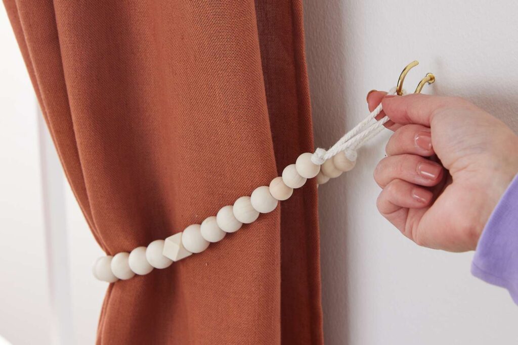 "Close-up of a hand adjusting a rust-colored curtain with a stylish wooden bead tieback, highlighting modern curtain design and elegant interior design."